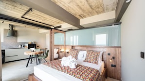 Een moderne hotelkamer met een houten hoofdbord, een groot bed met kleurrijke beddengoed, een klein bureau met stoel, en een keuken in de achtergrond.