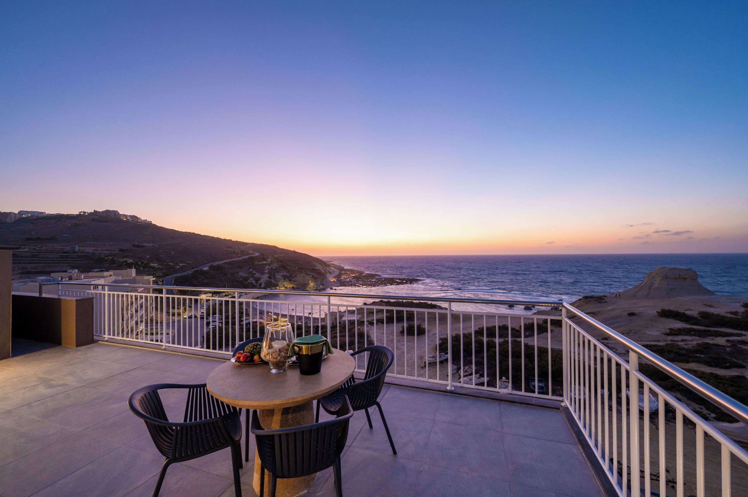 A balcony with a table and chairs overlooking a coastal landscape during sunset.
