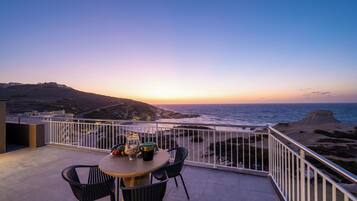 A balcony with a table and chairs overlooking a coastal landscape during sunset.