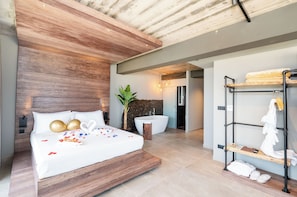 Ein modernes Hotelzimmer mit einem hölzernen Kopfteil, einem großen Bett mit weißen Leinen, einer weißen Badewanne und einem Holzregal mit Handtüchern.