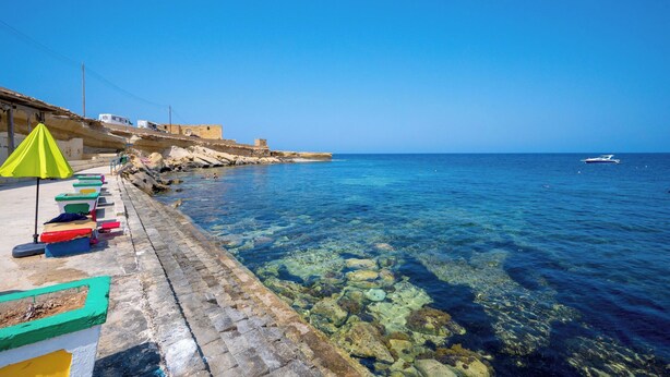 A coastal scene with clear blue water, a stone pier with colorful lounge chairs, and a boat in the distance.