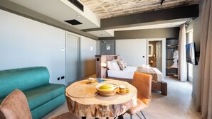 A modern hotel room with a green sofa, a round wooden table with breakfast items, a bed with a white comforter, and a TV mounted on the wall.