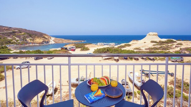 A balcony with a table set for two, overlooking a beach with a clear view of the sea and a distinctive rock formation. - Salini Suites Luxury Accommodation With Terrace and Sea View 101 (Iż-Żebbuġ)