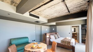 A modern hotel room with a wooden ceiling, a bed with a patterned blanket, a green sofa, and a round table with breakfast items. - Salini Suites Luxury Accommodation With Terrace and Sea View 101 (Iż-Żebbuġ)