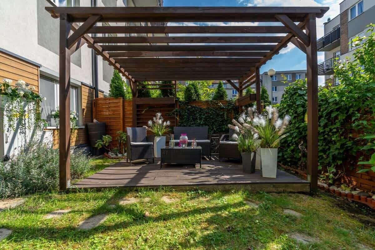 Una pérgola de madera con mobiliario exterior, un sendero y follaje.
