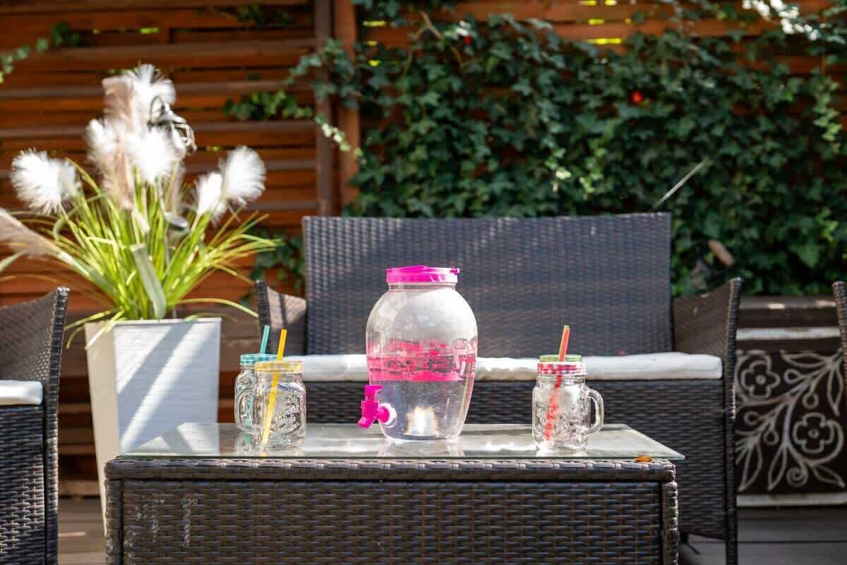 A wicker table with a glass jar and two small jars with straws, set outdoors with a wooden fence and greenery in the background.