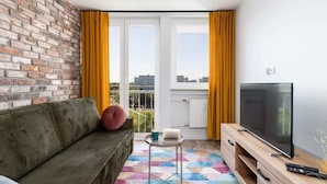 A living room with a brick wall, a sofa, a TV on a wooden stand, a small table with a book, and yellow curtains. - Elegant Apartment in Wroclaw | Close to Main Railway Station (Wroclaw)