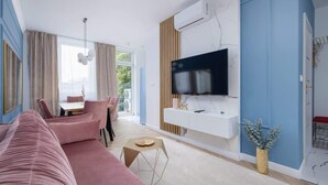 A modern living room with a pink sofa, a dining area with a table and chairs, a flat-screen TV mounted on a white wall, and a large window with curtains. - Elegant Apartment | Air Conditioning, Balcony, Close to Market Square (Kraków)