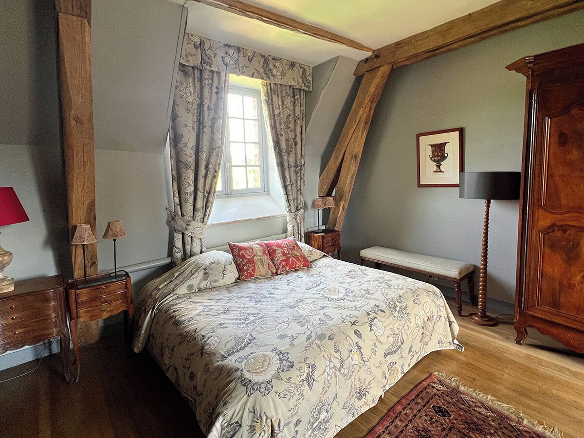 A bedroom with a large bed, wooden beams, a window with curtains, a bedside table with lamps, a bench, and a wooden cabinet.