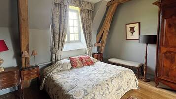 A bedroom with a large bed, wooden beams, a window with curtains, a bedside table with lamps, a bench, and a wooden cabinet.