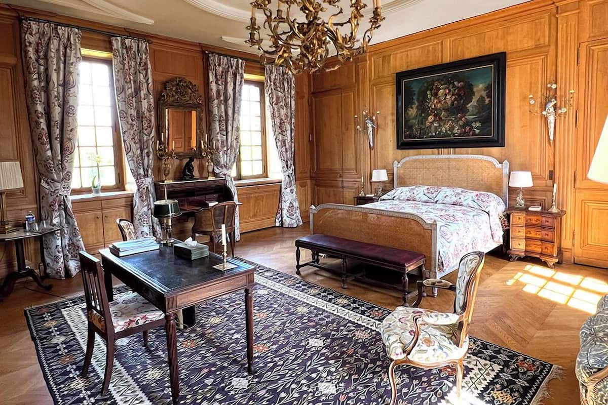 A bedroom with a large bed, a desk, a chair, a chandelier, and a painting on the wall.