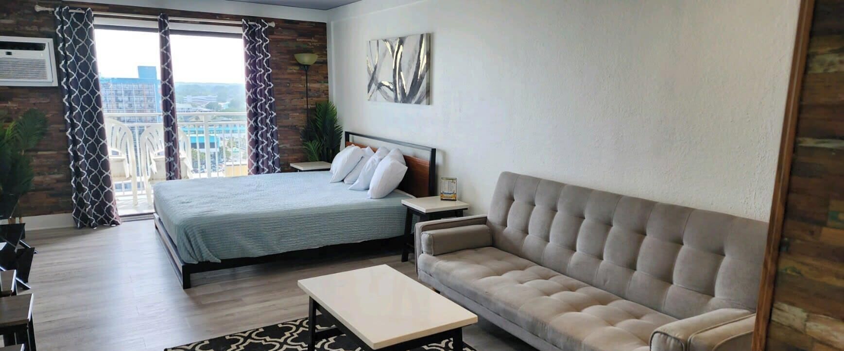 A modern hotel room with a large bed, a sofa, a coffee table, and a view of the city.