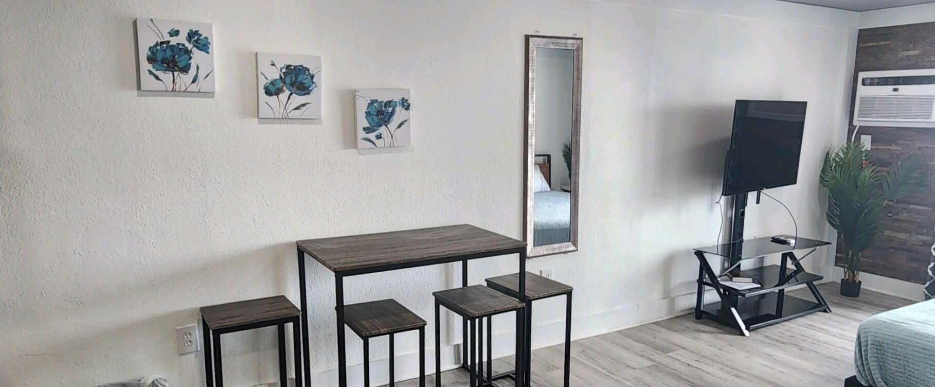 A room with a wooden table, two stools, a TV on a stand, and three floral paintings on the wall.