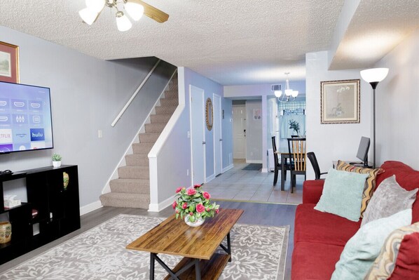 A living room with a staircase, a TV, a coffee table with flowers, a red sofa, and a dining area with chairs and a table. - 3-Bed Athens Getaway right near UGA! Pool, Parking, SmartTV (Athens)