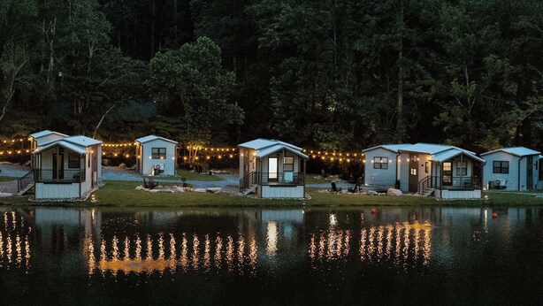 A row of small cabins by a lake with string lights, surrounded by trees.