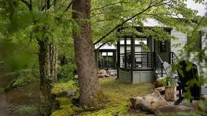 A cabin with a wooden deck, a covered porch, and a view of a forested area with a stream.