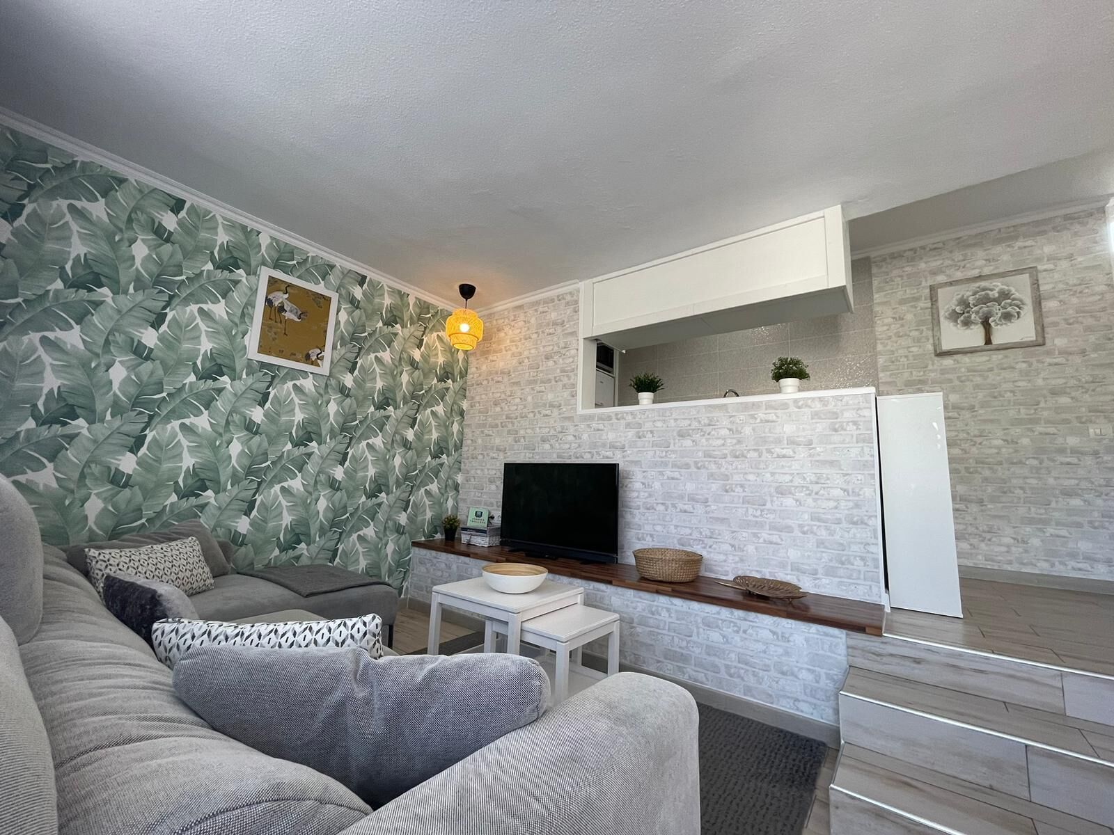 Ett modernt vardagsrum med en grå soffa, en platt-TV, ett vitt soffbord och en köksdel med ett vitt kylskåp och en väggbeklädnad av sten.