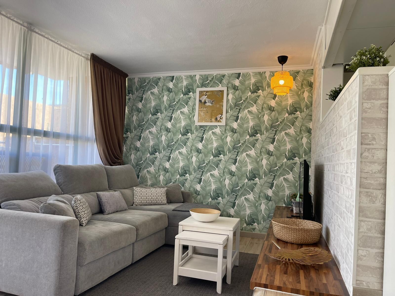 Un salon avec un canapé gris, une petite table blanche, un buffet en bois et un mur recouvert d’un papier peint à motifs de feuilles.