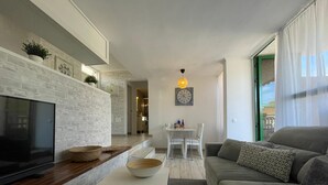 A modern living room with a flat-screen TV, a white brick wall, a dining area with a table and chairs, and a sofa with patterned cushions.