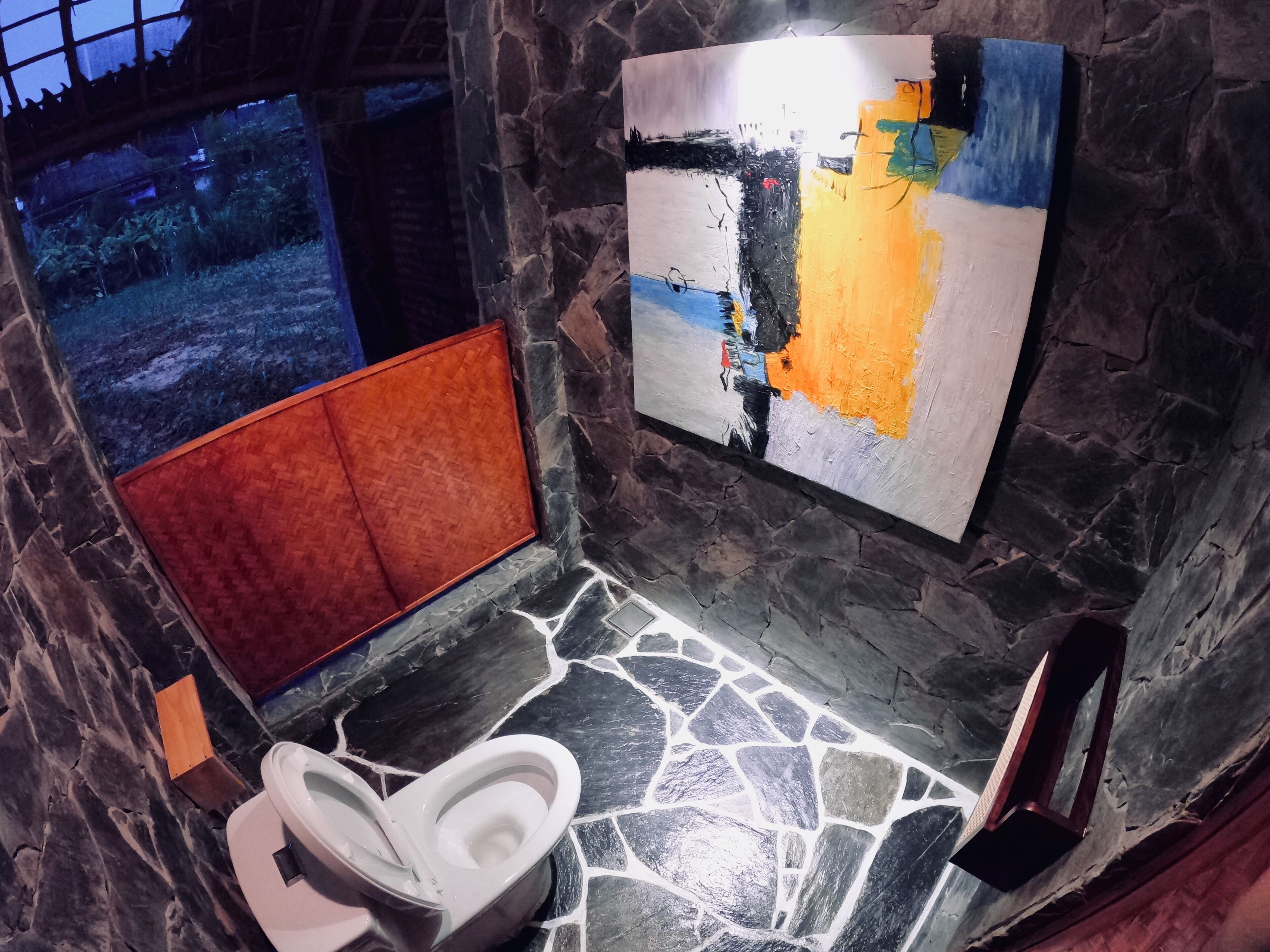 Un baño con piso de piedra, inodoro blanco, banca roja y un cuadro abstracto grande en la pared.