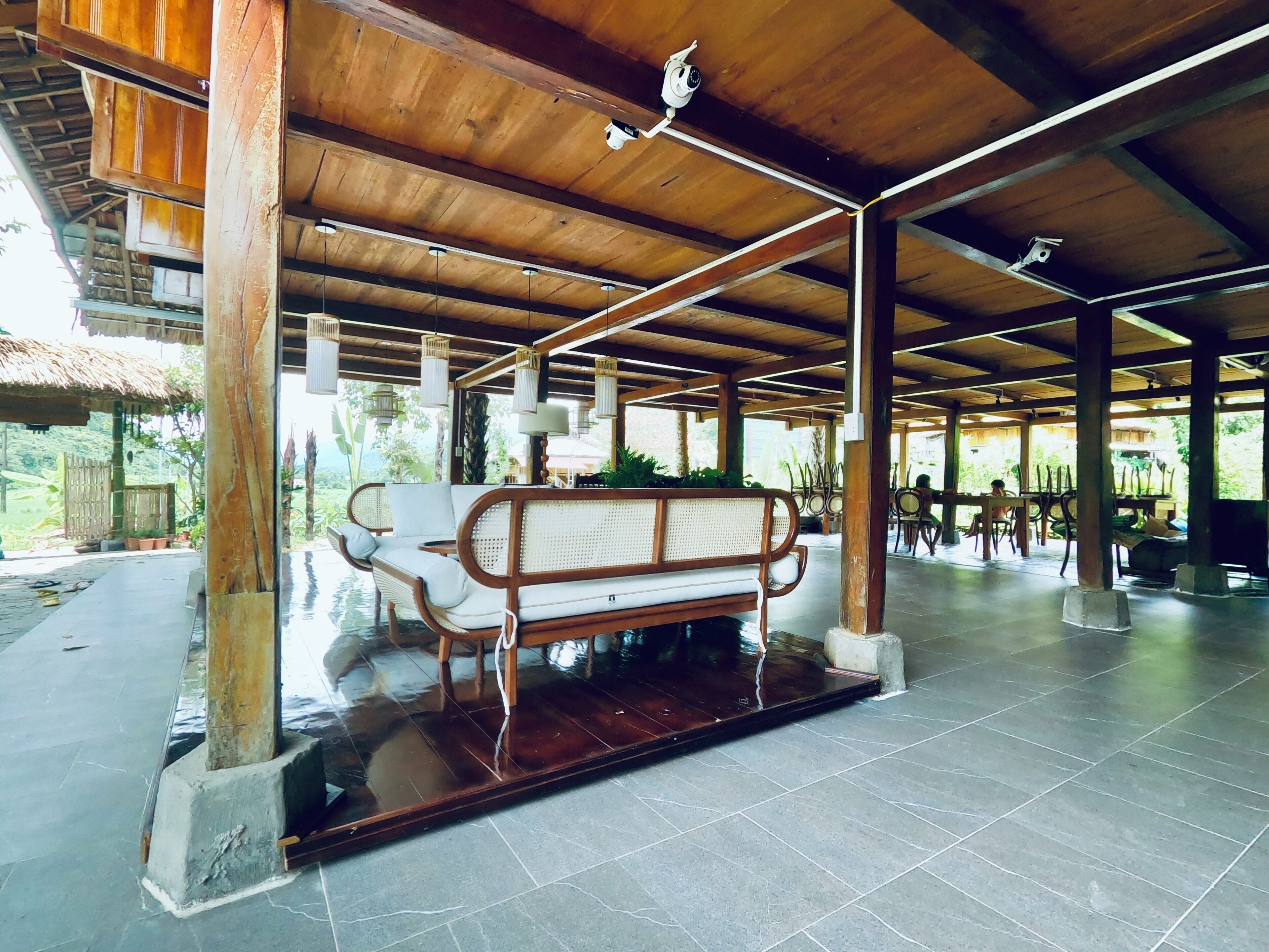 Un espacio al aire libre techado con un banco y mesas de madera, rodeado de pilares y un suelo de baldosas.