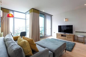 En moderne stue med sofa, skammel, fjernsyn og udsigt gennem store vinduer.