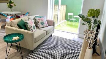 Un salon avec un canapé, une petite table ronde, un tapis à motifs et une vue sur un jardin verdoyant par une porte ouverte.