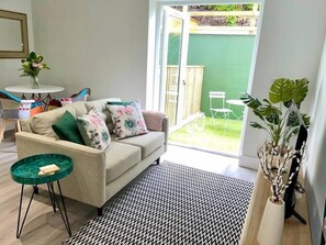 A living room with a sofa, a small round table, a patterned rug, and a view of a green garden through an open door. - Gorgeous New 1-Bed, Town Centre, Parking & Garden (Bournemouth)
