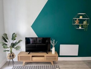 A modern living room with a flat-screen TV on a wooden console, a large potted plant, and a geometric wall design. - Gorgeous New 1-Bed, Town Centre, Parking & Garden (Bournemouth)