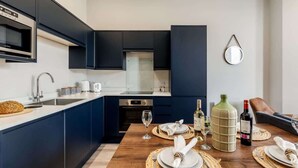 A modern kitchen with dark blue cabinets, a stainless steel oven, and a dining area set with a wooden table, plates, and wine glasses. - Gorgeous Family 2-Bed Apt w/ Free Gym & Parking (Bournemouth)