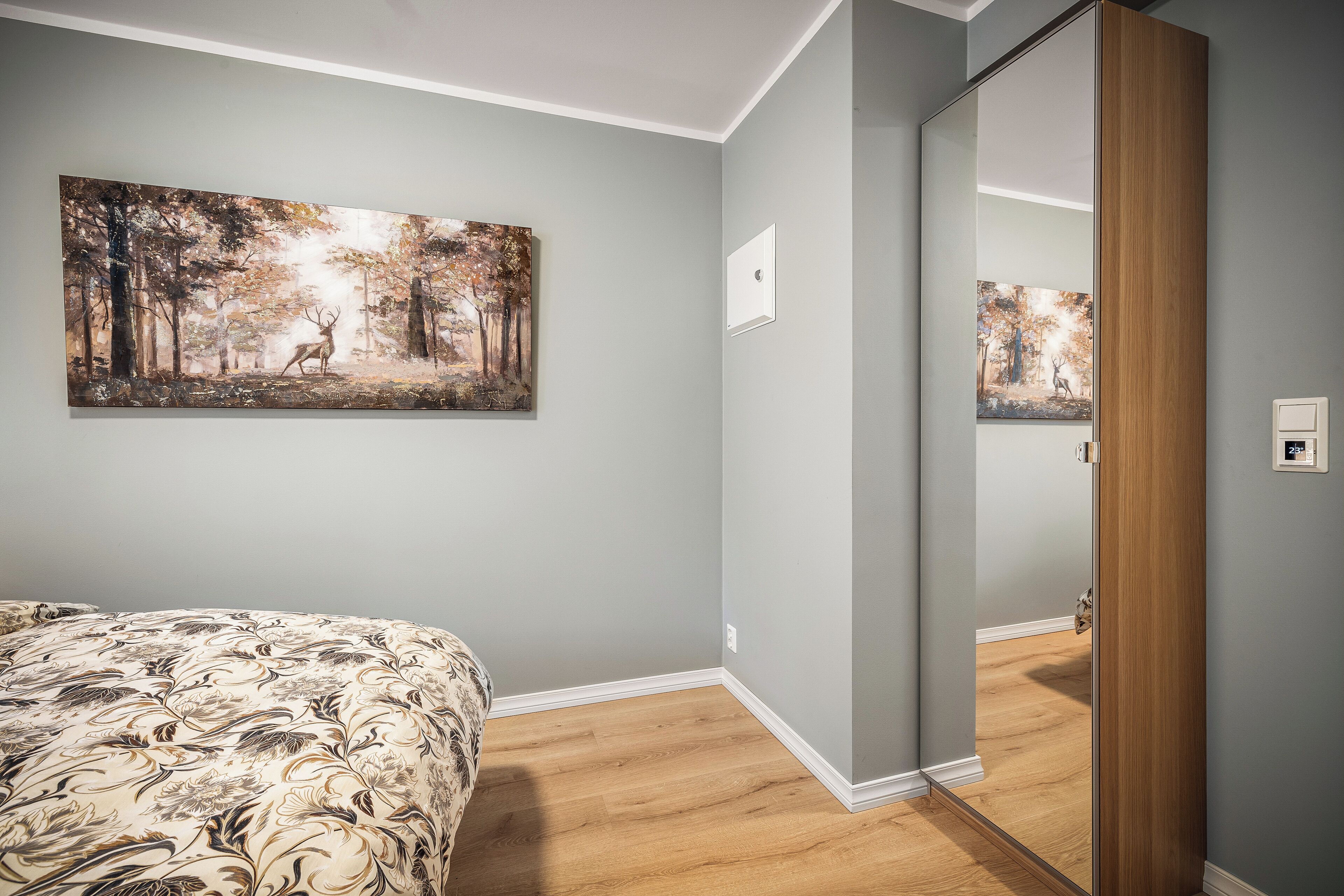A bedroom with a bed, a painting of a forest scene, and a wooden door leading to another room.
