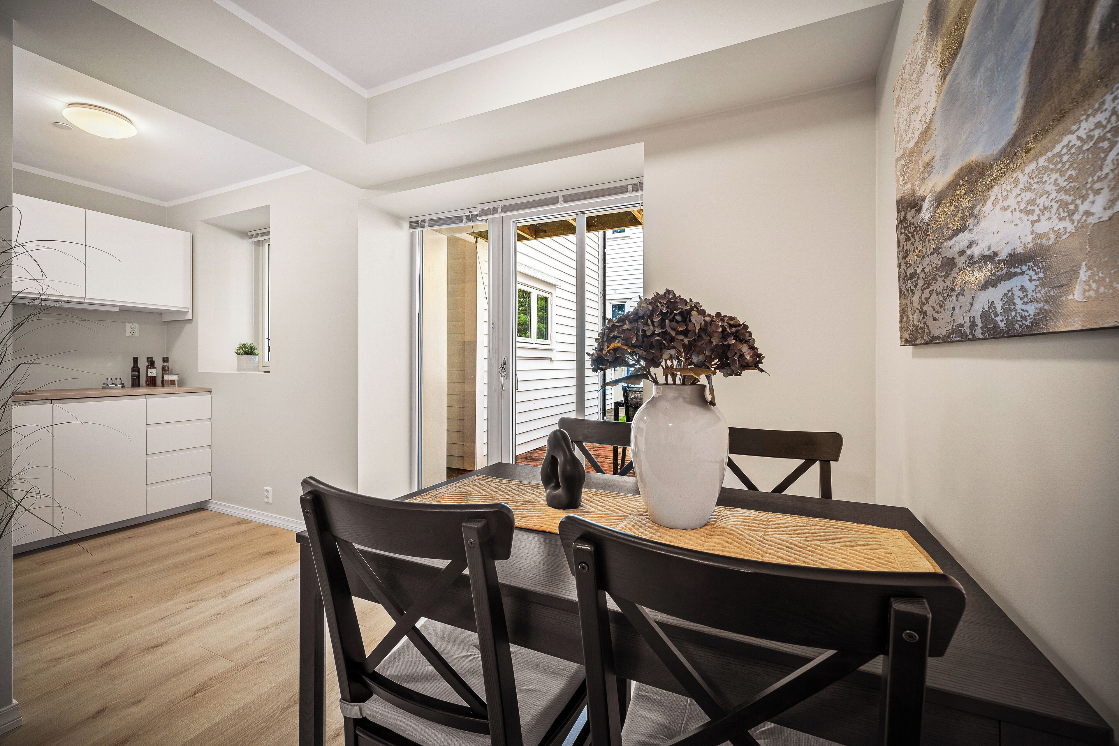 Un coin repas avec une table dressée pour quatre personnes, un vase contenant des fleurs séchées et un tableau accroché au mur.