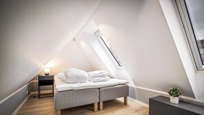 Ein modernes Schlafzimmer im Loftsstil mit schrägen Wänden, einem Bett mit grauem, gesteppten Kopfteil, einem Nachttisch mit Lampe und einem kleinen Topf mit Pflanze auf einer Konsole.