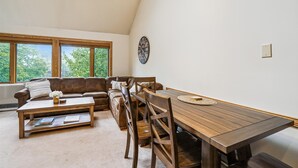 Una sala acogedora con un sofá de cuero marrón, una mesa de centro de madera y una mesa de comedor con sillas. Se ve una ventana grande con vistas a árboles, un reloj de pared y una lámpara colgante.