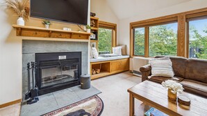 Una sala de estar acogedora con chimenea, un sofá cómodo, una mesa de centro y un ventanal con vistas a árboles.