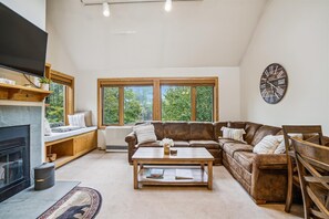 Un salon chaleureux avec une cheminée, un canapé en cuir marron, une table basse et une grande fenêtre donnant sur un paysage verdoyant.