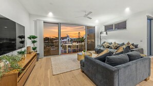 A modern living room with a grey sofa, a flat-screen TV, wooden flooring, and a view of a sunset through a glass door.