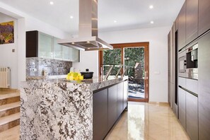 A modern kitchen with a marble island, stainless steel appliances, and a view of a garden through a glass door. - Villa Finca Estel (Calvia)
