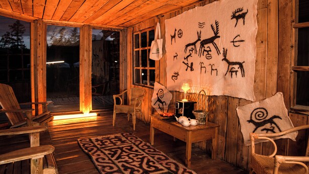 A wooden cabin interior with a large tapestry depicting animal drawings, a table with a lamp, chairs, and a patterned rug. - Nature Door Resort. Khuvsgul province. (Alag-Erdene)