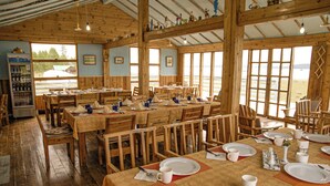 A rustic dining area with wooden tables and chairs, a beverage dispenser, and large windows offering a view of the outdoors. - Nature Door Resort. Khuvsgul province. (Alag-Erdene)
