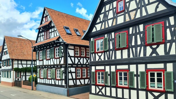 En række traditionelle bindingsværkshuse med røde og grønne skodder, en klar himmel og en belagt vej.