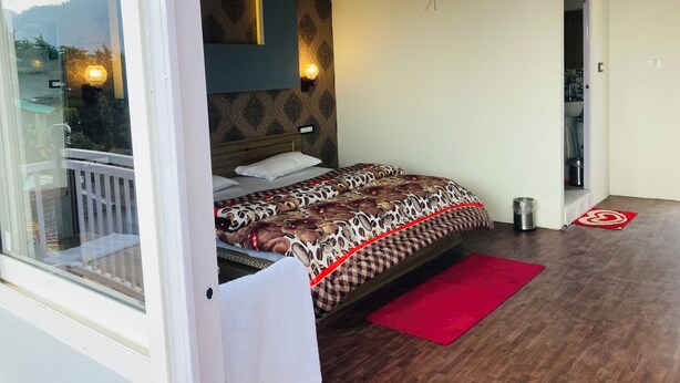 A bedroom with a bed, a red rug, a wooden floor, a patterned wall, and a balcony with a railing. - Sizzling Resort And Adventure (Dhanaulti)