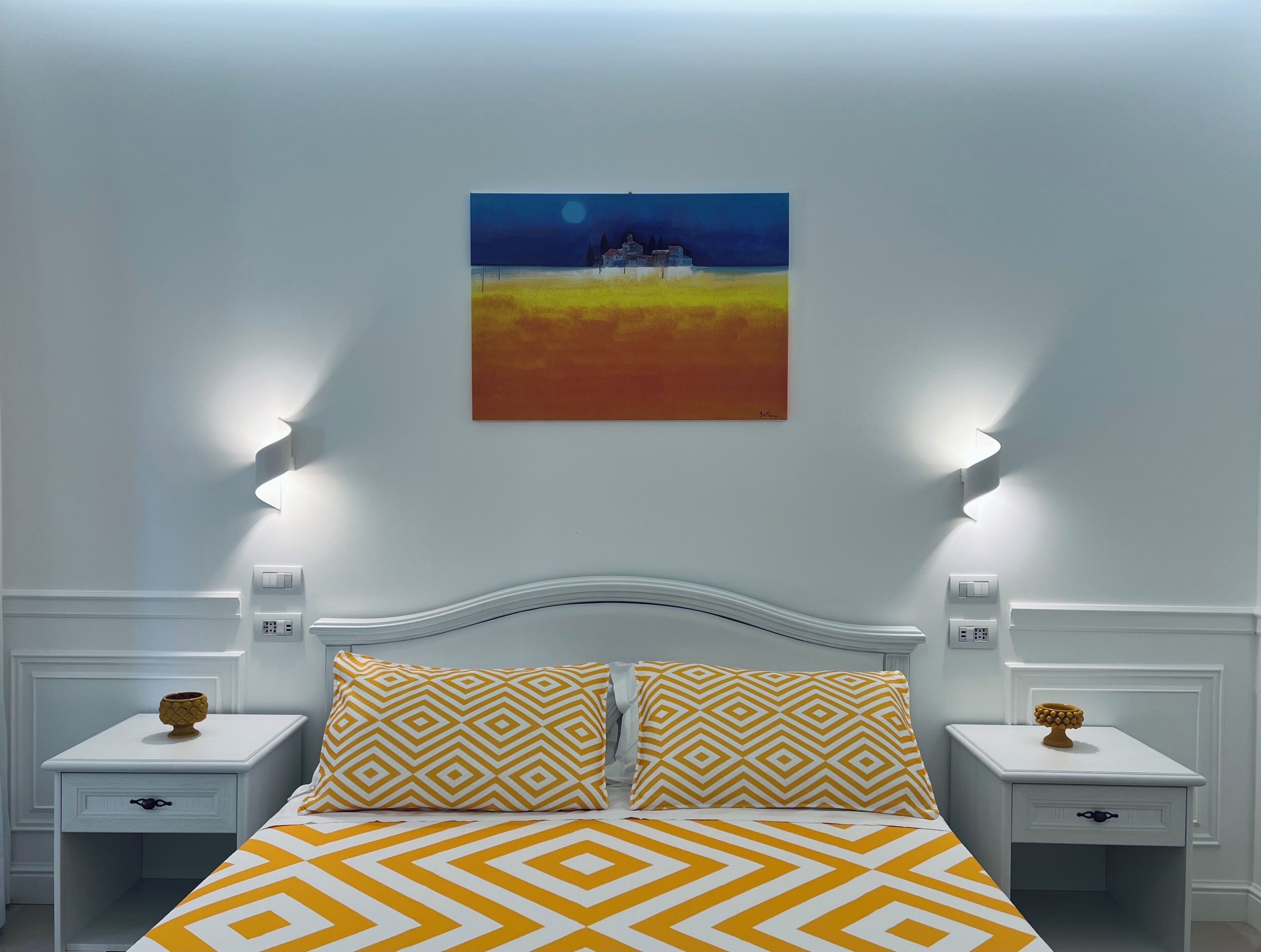 A bedroom with a bed, two nightstands, a painting, and wall-mounted lights.