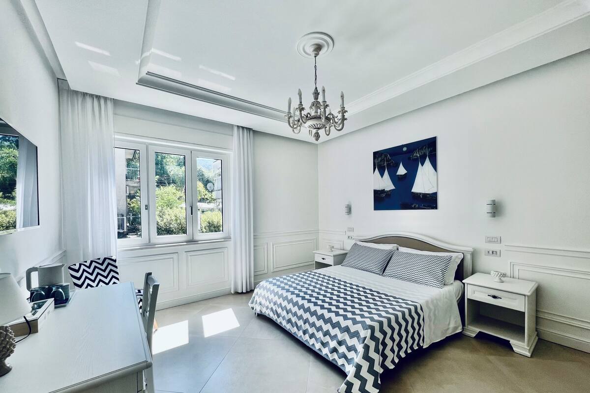 A bedroom with a bed, bedside tables, a chandelier, a window with curtains, and a painting on the wall.