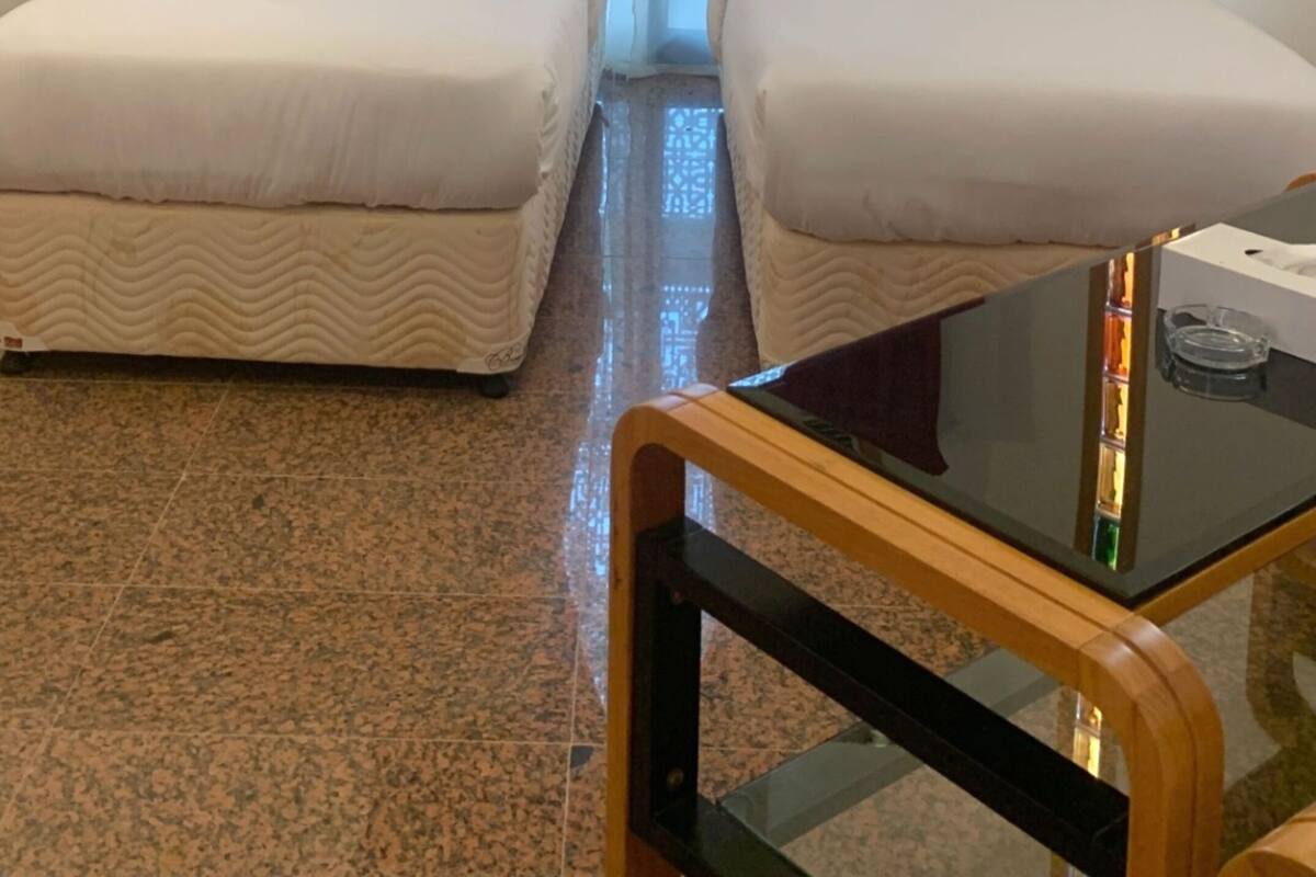 A hotel room with two single beds, a small table, a glass-top table, an air conditioning unit, and a window with red curtains.