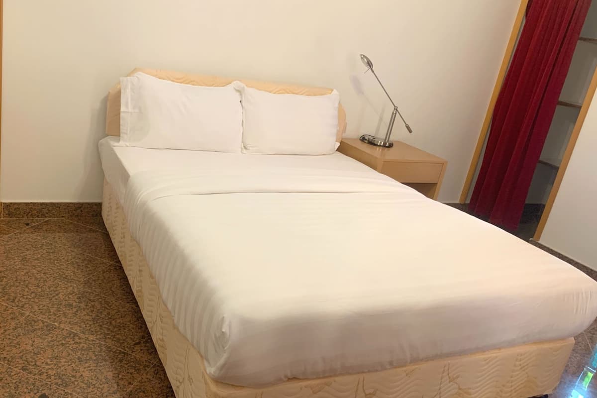 A neatly made bed with white linens in a room with a wooden door and a nightstand with a lamp.