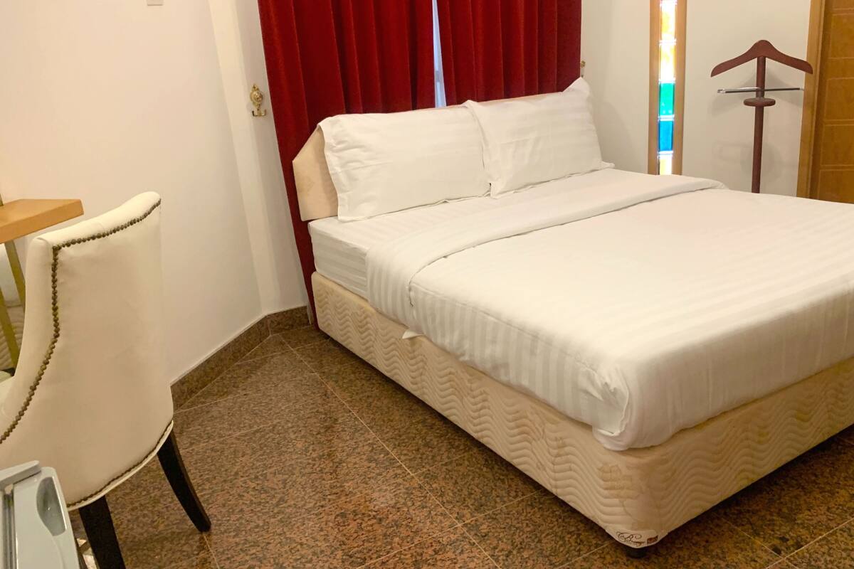 A neatly made bed with white linens in a room with a red curtain, a wooden door, and a small table with a chair.