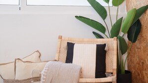 Ein Korbstuhl mit grau-weißem Kissen, eine große, grüne Pflanze im schwarzen Topf und ein gewebter Teppich auf Holzdielen.
