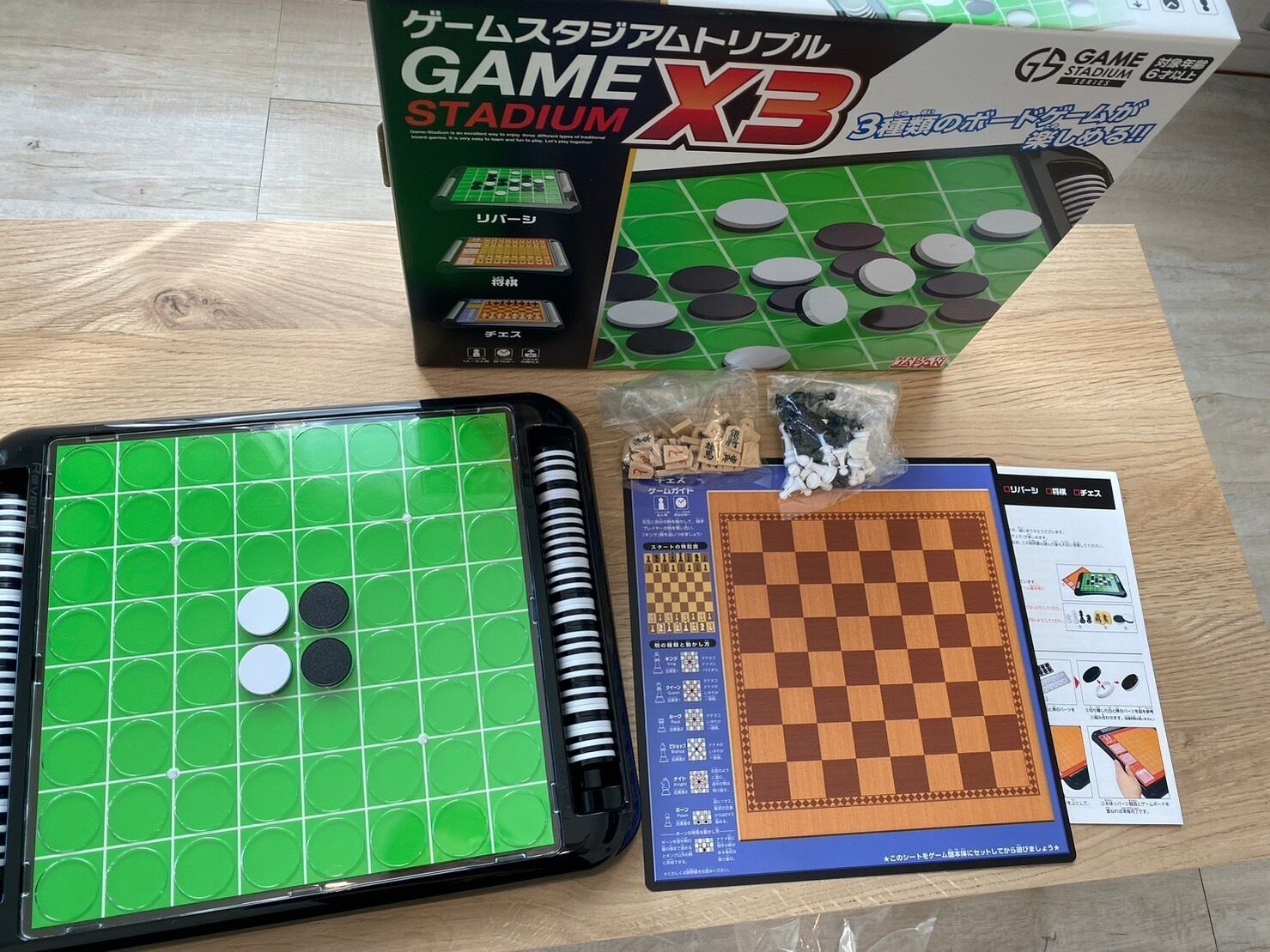 A GAME X3 STADIUM chess set with a green playing board, pieces, and instruction booklet on a wooden table.