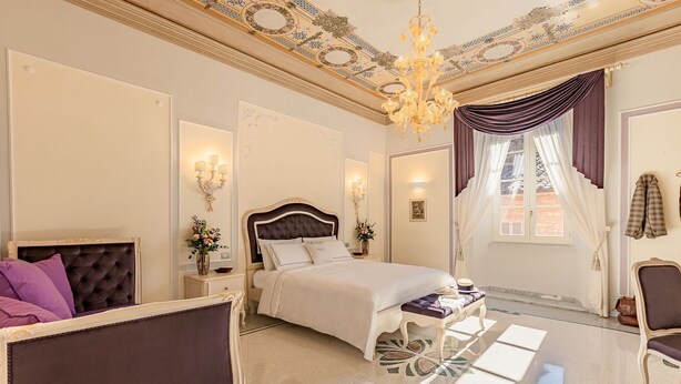A luxurious bedroom with a large bed, a chaise lounge, a chandelier, and a decorative ceiling.